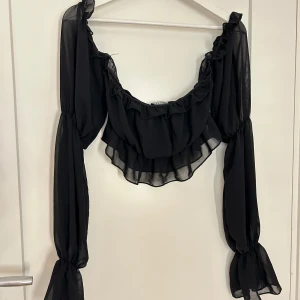 Svart offshoulder blus med volang - Säljer en svart offshoulder blus i transparent material med volangdetaljer längs halsringningen och långa puffiga ärmar med knyt vid handlederna. Perfekt för dig som vill ha en trendig och luftig topp.