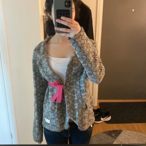 Grå mönstrad stickad cardigan med rosa knytband - Mysig grå stickad cardigan med vitt mönster och ett rosa knytband i midjan. Jackan har lång ärm, bred krage och broderad detalj på ärmen. Perfekt för lager-på-lager och en avslappnad stil.