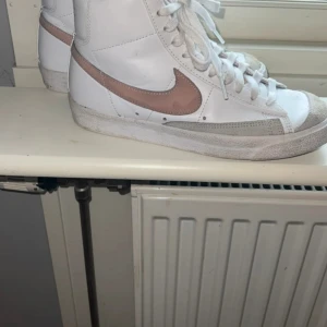 Nike Blazer Mid '77 i vitt och beige - Säljer ett par Nike Blazer Mid '77 i vitt skinn med rosa swoosh och detaljer. Klassisk hög modell med snörning och vit platt sula. Perfekta för dig som gillar retrostil och vill ha något som sticker ut lite extra.