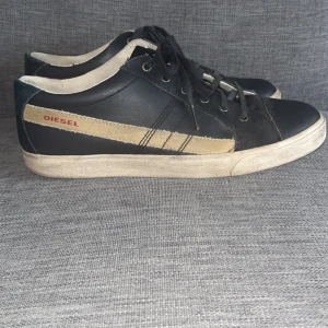 Svarta sneakers från Diesel - Säljer ett par ovanliga svarta sneakers från Diesel med beige ränder på sidorna och logga i rött. Skorna har snörning och rund tå, samt vit platt sula. Perfekta för en avslappnad stil. Tar emot prisförslag