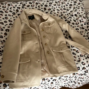 Beige jacka från Dorothy Perkins - Stilren beige jacka från Dorothy Perkins med fyra fickor framtill, knappar och bälte i midjan. Jackan har lång ärm och klassisk krage, perfekt för en snygg och enkel look.