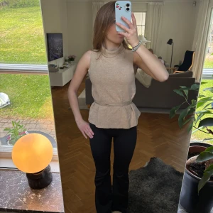 Beige ärmlös tröja med polokrage - Säljer en beige ärmlös topp med polokrage och peplumvolang i midjan. Toppen har en markerad midja och är i ett mjukt material.