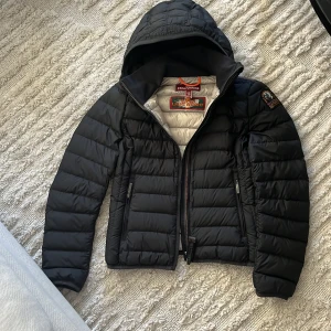 Svart dunjacka med huva från Parajumpers ”BARN” - Säljer en svart, superlätt dunjacka från Parajumpers med huva. Jackan har quiltad design, dragkedja framtill och två fickor med dragkedja. Insidan är ljusgrå och jackan har en patch på ena ärmen.