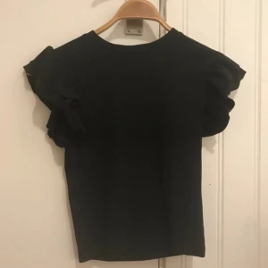 Svart topp med volangärmar - Säljer en svart topp med korta volangärmar. Enkel och stilren design som passar perfekt till många olika outfits. Toppen har rund halsringning och är gjord i ett mjukt material.