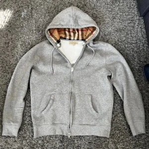  Burberry kofta  - Snygg grå hoodie från Burberry med klassiskt rutigt mönster i huvan. Perfekt för en stilren och avslappnad look. Den har dragkedja framtill och praktiska fickor.