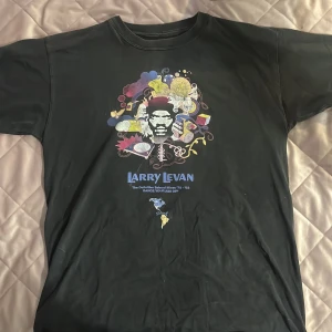 Svart band t-shirt med Larry Levan - Svart bandtröja med färgglatt tryck av Larry Levan och olika grafiska element på bröstet. Texten 'Larry Levan' syns tydligt under motivet. Klassisk passform och rund halsringning. Perfekt för dig som gillar musik och streetwear.