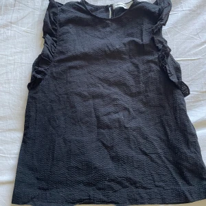 Svart ärmlös topp med volangdetaljer - Svart ärmlös topp med volanger längs axlarna och sidorna. Toppen har en lätt strukturerad yta och rund halsringning. Perfekt för att lyfta en enkel outfit.