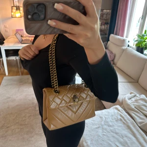 Kurt geiger väska - Säljer en beige/orange quiltad axelväska från Kurt Geiger med guldfärgad kedja och snyggt emblem framtill. Väskan har ett klassiskt lock och rymligt fack inuti. Perfekt för dig som vill ha en stilren och trendig accessoar.