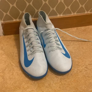 Vita och blåa Nike fotbollsskor - Säljer ett par vita Nike fotbollsskor med blå detaljer och stor Nike-logga på sidan. Skorna har snörning och platt sula, perfekt för inomhusspel. Skorna kommer också med paket. Skorna är köpta för 850. Snygg och sportig design med högre skaft för extra stöd. Pris går att diskutera ifall man inte är nöjd med det nuvarande priset