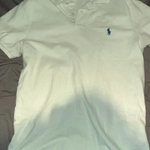Vit pikétröja från Polo Ralph Lauren - Vit pikétröja från Polo Ralph Lauren med klassisk krage och två knappar. Tröjan har korta ärmar och blå broderad logga på bröstet. Modellen är slim fit och passar perfekt till en stilren look. Priset kan diskuteras!