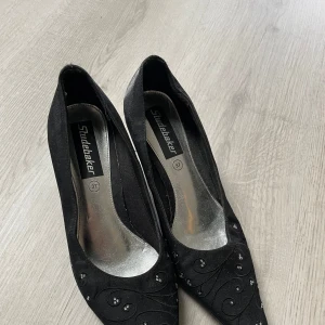 Svarta pumps med broderi och pärlor från Studebaker - Säljer ett par eleganta svarta pumps från Studebaker i storlek 37. Skorna har broderade detaljer och små pärlor på ovandelen samt en klassisk klack. Perfekta för dig som vill ha något stilrent med lite extra detalj.