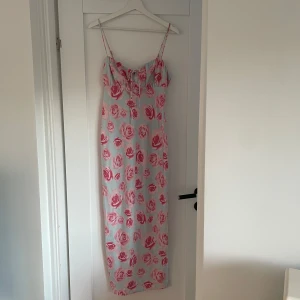 Ljusblå långklänning med rosa rosor - Säljer en ljusblå långklänning med rosa blommigt mönster och smala axelband. Klänningen har en söt knytdetalj framtill och figurnära passform. Perfekt för sommarens alla tillfällen!
