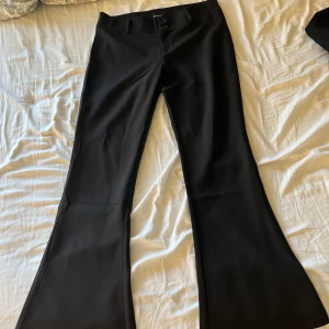 Svarta bootcut kostymbyxor från Gina Tricot - Säljer ett par svarta kostymbyxor från Gina Tricot i bootcut-modell. Byxorna har hög midja, vida ben och knäppning fram samt fickor med lock och knapp bak. Perfekta för en stilren look.