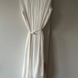 Vit ärmlös långklänning med knytband - En stilren vit långklänning med ärmlös design och rund halsringning. Klänningen har ett knytband i midjan som ger en snygg siluett. Perfekt för sommarens alla tillfällen.