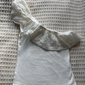 Vit oneshoulder topp med spets från Holly & White - Säljer en vit oneshoulder topp från Holly & White med bred volang i spets och broderade blommor. Perfekt för dig som vill ha något unikt och somrigt med fina detaljer.