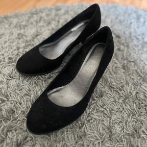 Svarta pumps med klack - Säljer ett par klassiska svarta pumps med rund tå och klack. Skorna har en stilren design och är tillverkade i mocka. Perfekta för dig som vill ha ett par enkla men snygga skor till garderoben.