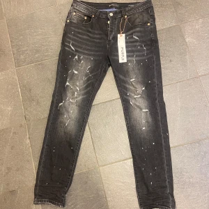 Svarta purple brand jeans - Säljer ett par svarta jeans från Purple Brand med coola slitningar och vita färgstänk. Klassisk femficksmodell med knappgylf och raka ben. Perfekta för dig som gillar en edgy streetstil. Ej autentisk 