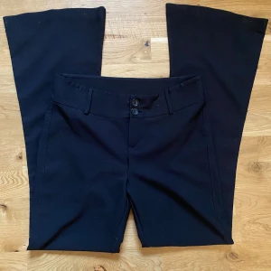 Svarta kostymbyxor från Amelie & Me - Svarta kostymbyxor från Amelie & Me med raka, vida ben och två knappar framtill. Byxorna har fejk-fickor bak och bälteshällor i midjan. Perfekta för en dressad look.