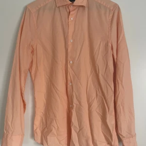 Persikofärgad skjorta - Riley - Säljer en stilren persikofärgad skjorta med klassisk krage och vita knappar. Skjortan har lång ärm och är perfekt för dig som vill ha en fräsch och enkel look. Passar lika bra till jeans som till kostymbyxor.