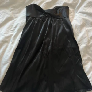 Svart axelbandslös satin klänning - Säljer en svart axelbandslös klänning i glansigt satinmaterial. Klänningen har en snygg twist-detalj vid bysten och en lätt utställd passform. Perfekt för dig som vill ha en stilren och elegant look.