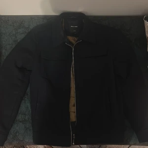 Jack & Jones Jacka - Säljer en svart jacka från Jack & Jones med dragkedja framtill och två bröstfickor. Jackan har lång ärm och en stilren design, perfekt för dig som gillar enkel och klassisk stil.