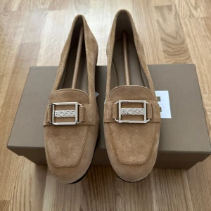 Beige mockaloafers från BOSS -  ett par stilrena beige loafers från BOSS i mocka. Skorna har en fyrkantig tå och en dekorativ metallspänne framtill med BOSS-logga. Platt sula och klassisk design som passar till många outfits.