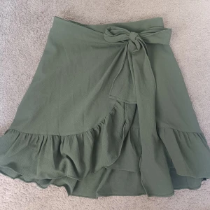 Grön omlottkjol med volang och knytband - Säljer en ljusgrön omlottkjol med volangkant och brett knytband i midjan. Kjolen är perfekt för en avslappnad och trendig look. Passar perfekt nu inför våren och sommaren. I superfint skick och är enkel att styla