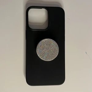 Svart mobilskal med glittrig popsocket - Säljer ett svart mobilskal med en stor, rund popsocket täckt av glittrande stenar. Popsocketen är vit undertill och ger ett extra grepp om mobilen. Perfekt för dig som vill ha lite extra bling på din telefon!