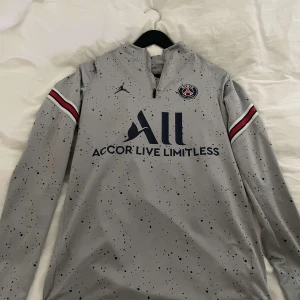 Grå PSG fotbollströja med Jordan-logga - Säljer en grå långärmad PSG fotbollströja med mörkblå och röda detaljer på ärmarna. Tröjan har Jordan-logga, klubbmärke och texten 'Accor Live Limitless' på bröstet. Unik stänkmönstrad design och dragkedja vid halsen.