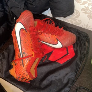 Röda Nike fotbollsskor med snörning - Säljer ett par röda Nike fotbollsskor med vit swoosh och svarta detaljer. Skorna har snörning och är tillverkade i syntetmaterial med mönstrad ovandel och plös. Perfekta för dig som vill sticka ut på planen! Och jag säljer dom på grund av att de är små för mig