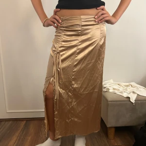 Champagnefärgad satinkjol från Cider - Säljer en lång, champagnefärgad satinkjol från Cider. Kjolen har snygg snörning och slits på sidan som ger en trendig look. Materialet är glansigt och känns lyxigt. Perfekt för dig som vill sticka ut lite extra.
