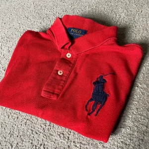 Röd pikétröja från Polo Ralph Lauren - Röd pikétröja från Polo Ralph Lauren ⚠️I storlek L junior (motsvarar storlek S⚠️) med klassisk krage, knäppning med två knappar och stor mörkblå broderad logga på bröstet. På vänster ärm finns en mörkblå siffra 3. Tröjan är i bomull och har korta ärmar, perfekt för en sportig och stilren look.