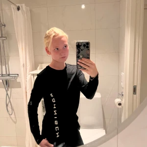 Svart långärmad tröja från Röhnisch - Svart långärmad tröja från Röhnisch med vit logga tryckt vertikalt längs ena sidan framtill. Tröjan har en clean och sportig look med rund halsringning och smal passform. Perfekt för dig som gillar stilrena och funktionella plagg.