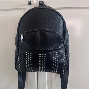 Svart ryggsäck med nitar och dragkedja - Cool svart ryggsäck i syntetmaterial med silvriga nitar framtill i rader. Väskan har en rundad form, dragkedja upptill och ett extra fack med dragkedja på framsidan. Perfekt för dig som vill ha en edgy och praktisk väska till vardagen.