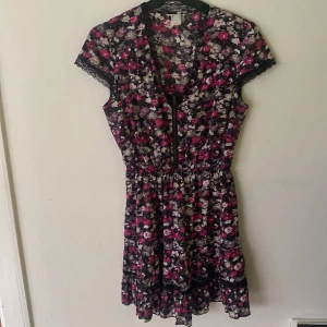 Blommig kortklänning med dragkedja - Kort bommulsklänning med blommigt mönster i svart, rosa, beige, grått och vitt. Klänningen har korta ärmar med spetsdetaljer, resår i midja och en dragkedja framtill. Perfekt för dig som gillar en feminin och lekfull stil!