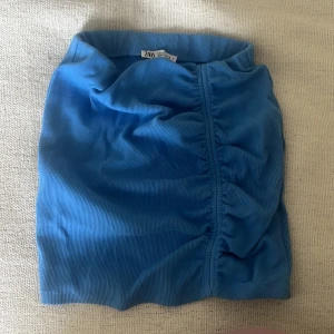 Blå kortkjol med rynk från Zara - Supersnygg blå kortkjol från Zara med rynkdetalj längs ena sidan. Kjolen har en tight passform och är gjord i ett ribbat material som ger en cool struktur. Perfekt att styla med en crop top eller oversized hoodie för en trendig look. Aldrig använd 