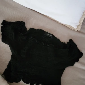 Svart smockad topp från Parisian - Supersöt svart smockad topp från Parisian med volangdetaljer och offshoulder-ärmar. Toppen är croppad och har en tight passform som framhäver midjan. Perfekt att styla med höga jeans eller kjol för en trendig look.