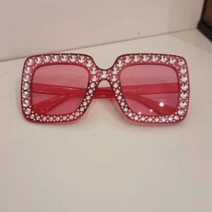 Rosa fyrkantiga solglasögon med strass - Stora fyrkantiga solglasögon i rosa med glittrande strass runt hela bågen. Glasen är tonade i rosa och bågarna är i plast. Perfekta för dig som vill sticka ut och addera extra bling till din stil.