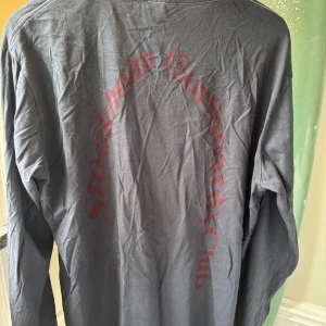 Svart långärmad tröja Stockholm Surfboard Club - Svart långärmad tröja från Stockholm Surfboard Club i 100% ekologisk bomull. Stor röd text i båge på ryggen. Snygga vita kontrastsömmar vid halsen. Perfekt för dig som gillar streetwear och vill ha något unikt.