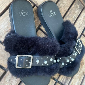 Svarta sandaler med päls och pärldetaljer - Coola svarta sandaler från VOX med fluffig fuskpäls och breda remmar i skinnlook. Remmarna har silvriga spännen och är dekorerade med pärlor och nitar för en edgy vibe. Perfekta för dig som vill sticka ut i sommar!