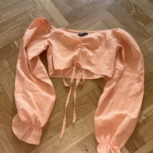 Persikofärgad croppad blus från SHEIN - Superfin persikofärgad croppad blus från SHEIN med långa puffärmar och knyt framtill. Tunt, lätt material med diskret rutstruktur och snygg rynkdetalj vid bysten. Perfekt för dig som vill sticka ut med en söt och trendig topp!