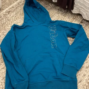 Blå hoodie från Wilson med broderi - Säljer en blå hoodie från Wilson med färgglatt broderat logotyp på bröstet. Tröjan har huva, magficka och långa ärmar. 