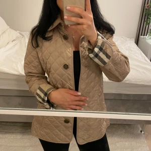 Beige quiltad jacka från Burberry XS (äkta) - Säljer en klassisk beige quiltad jacka från Burberry i storlek XS. Jackan har rutigt foder, stora knappar och snygga detaljer vid ärmsluten. 