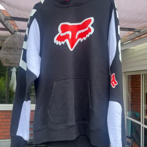 Svart och vit hoodie från Fox Racing - Säljer en svart och vit hoodie från Fox Racing med rött logotryck på bröstet och ärmen. Tröjan har huva, känguruficka och långa ärmar med vita detaljer. Crossmärke.