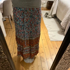 Blommig långkjol med volang - Säljer en färgglad långkjol med blommigt mönster i blått, rött och orange. Kjolen har volang nertill och knytband i midjan för en skön passform. Perfekt för dig som gillar boho-stil och vill ha något unikt i garderoben.
