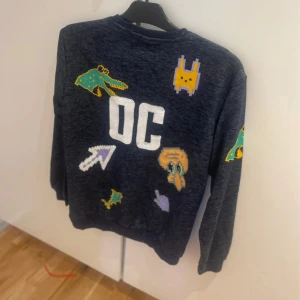 Mörkblå sweatshirt med pixeltryck Al-Safa strl M  - Cool mörkblå sweatshirt från Al-Safa med stora vita bokstäver på bröstet och färgglada pixelgrafik-tryck som krokodil, kanin och pilar. Tröjan har rund halsringning och långa ärmar. Perfekt för dig som gillar unika och lekfulla plagg.