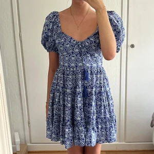 Blommig blå kortklänning från Zara - Supersöt blå kortklänning från Zara med vitt blommönster, puffärmar och smockad rygg. Klänningen har v-ringning, volangig kjol och knytband med tofsar framtill. Använd en gång. 