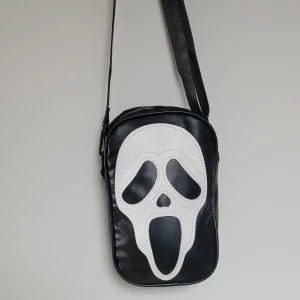  Helt ny Svart axelväska med Scream-motiv - Cool svart axelväska i syntet med stort vitt Scream-ansikte på framsidan. Väskan har justerbar rem och dragkedja upptill. Perfekt för dig som gillar skräckfilmer och vill sticka ut med din stil.