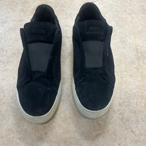 Svarta sneakers från Axel Arigato - Stilrena svarta sneakers från Axel Arigato i mocka med bred resår över vristen och vit platt sula. Minimalistisk design med diskret logga i guld på sidan. Perfekta för dig som gillar en clean och modern look.