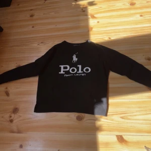 Svart långärmad tröja Polo Ralph Lauren - Svart långärmad tröja från Polo Ralph Lauren med stor vit logga och text på bröstet. Klassisk rak passform och rund halsringning. Perfekt för dig som gillar stilrena och ikoniska plagg med tydlig branding.